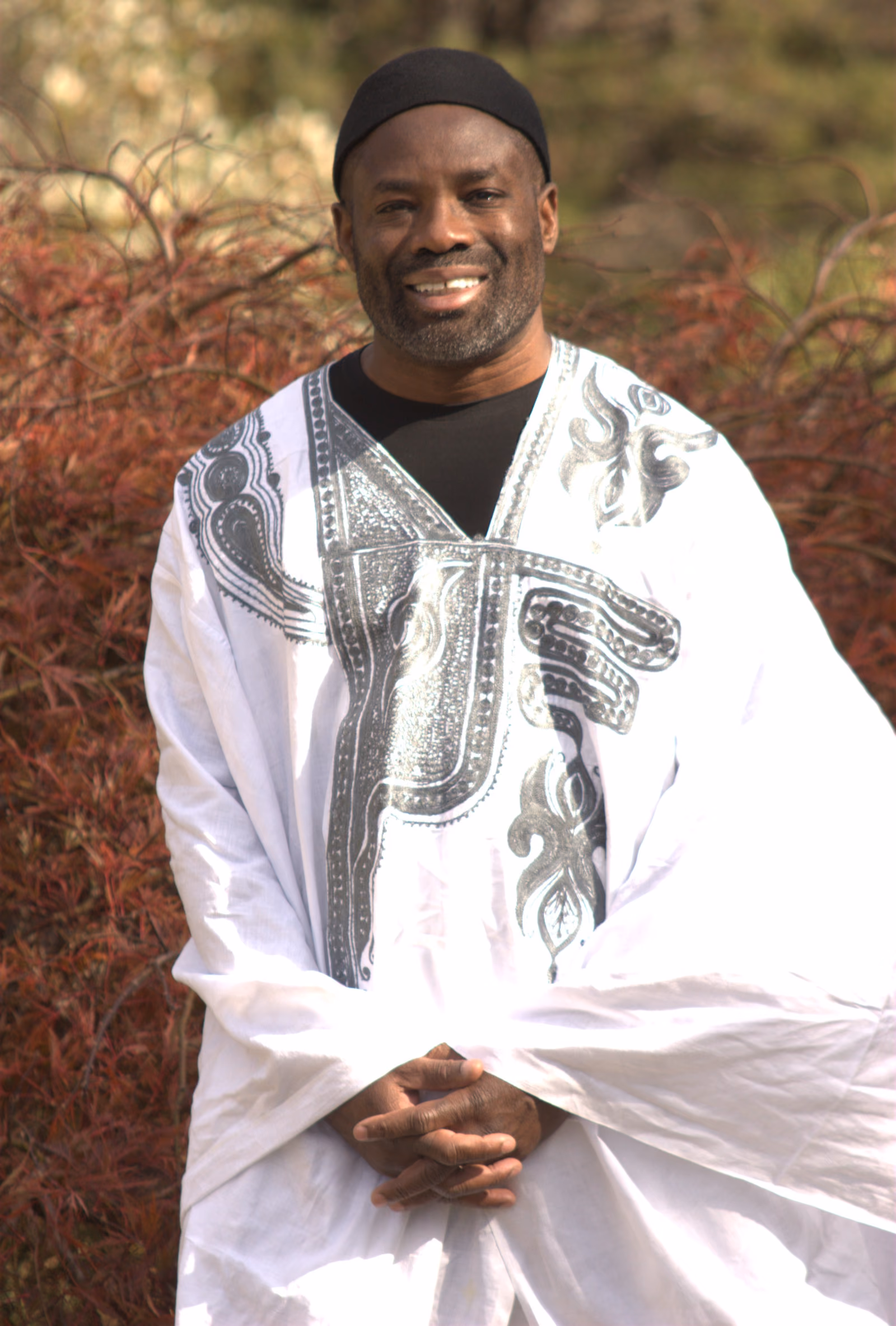 A man stands in a wide-sleeved white agbada, its chest and shoulders worked in dark embroidery, over a matching cap.
