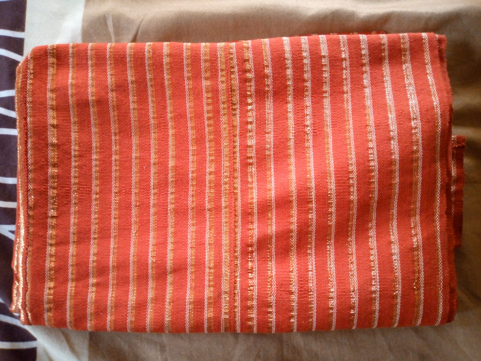 A length of coral-red aso oke folded on a plain surface, its narrow woven stripes and slight sheen running across the fold.