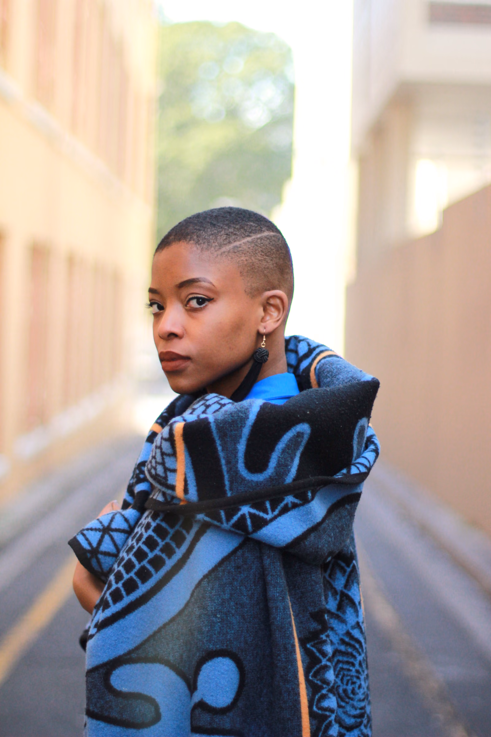 A young Mosotho woman wraps a blue Seanamarena blanket around her shoulders, its black geometric motifs clearly visible, standing in a sunlit street.