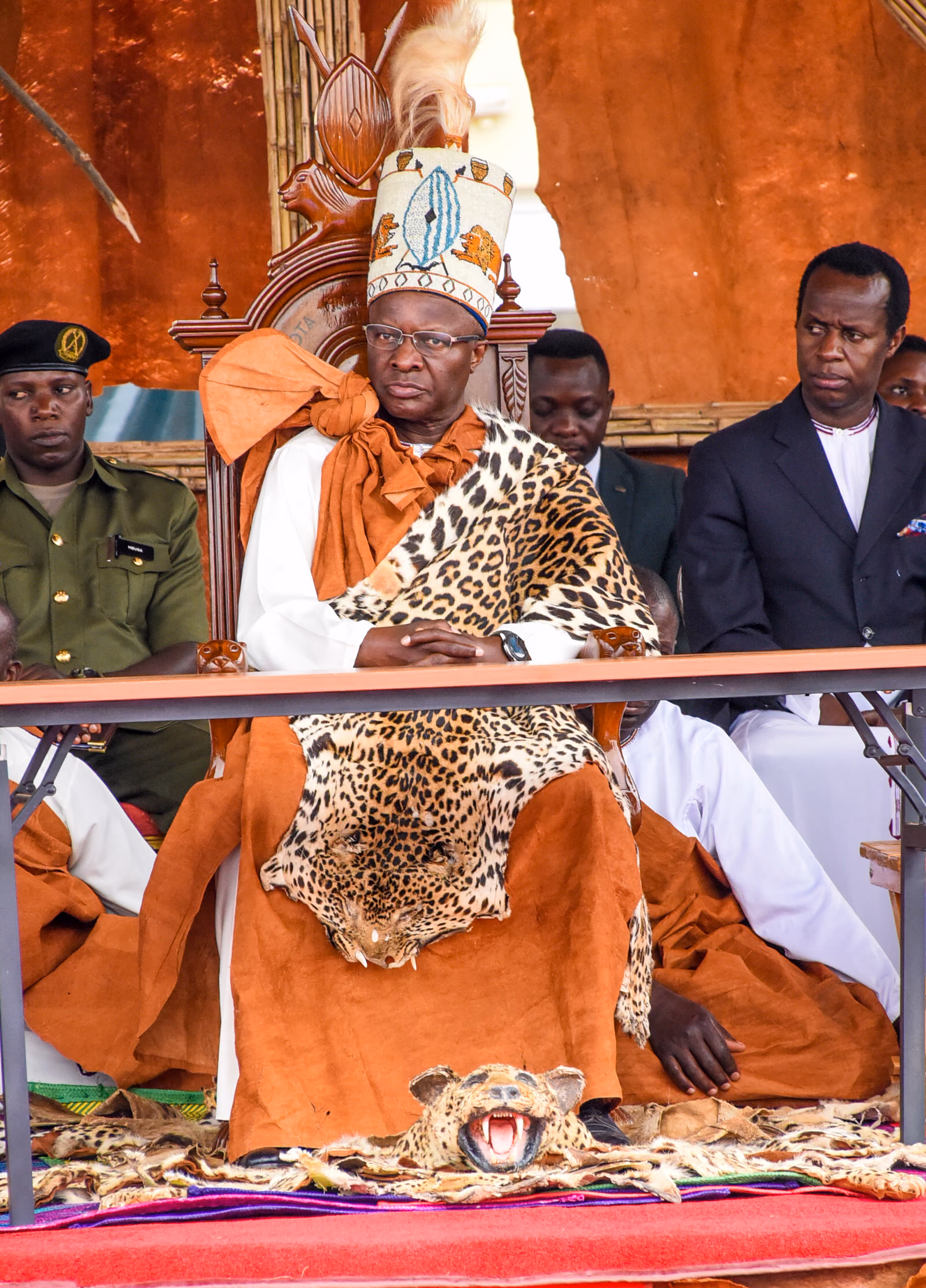 A seated ruler in orange ceremonial dress with leopard skin across the shoulders and a feathered hat, attendants standing behind.