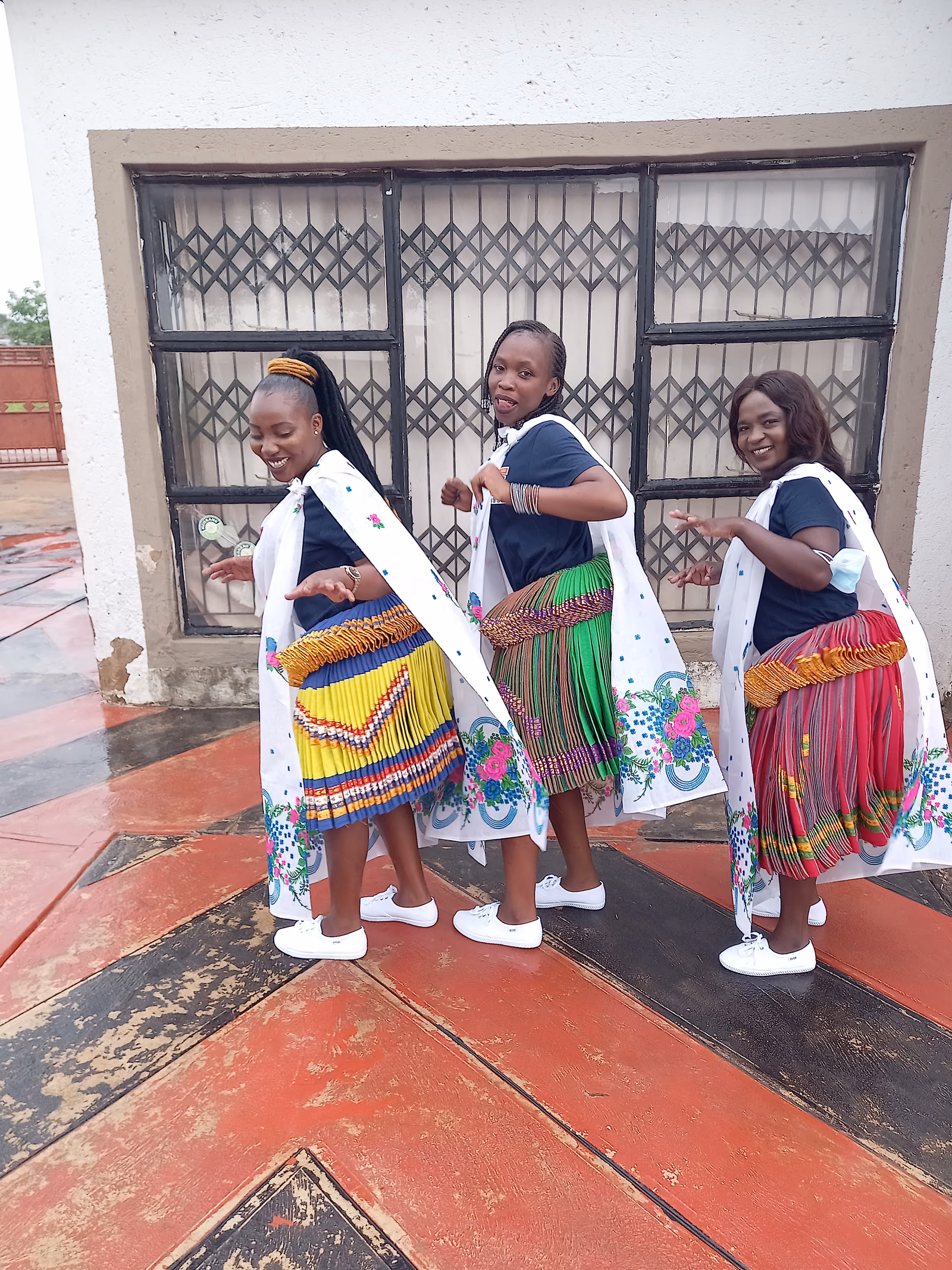 Three women stand together in short, densely pleated xibelani skirts in bright colours, with beaded tops and headwraps.