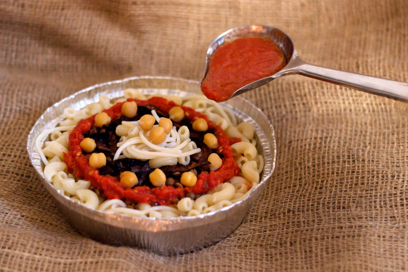A bowl of koshari layered with rice, lentils, pasta and chickpeas under tomato sauce.