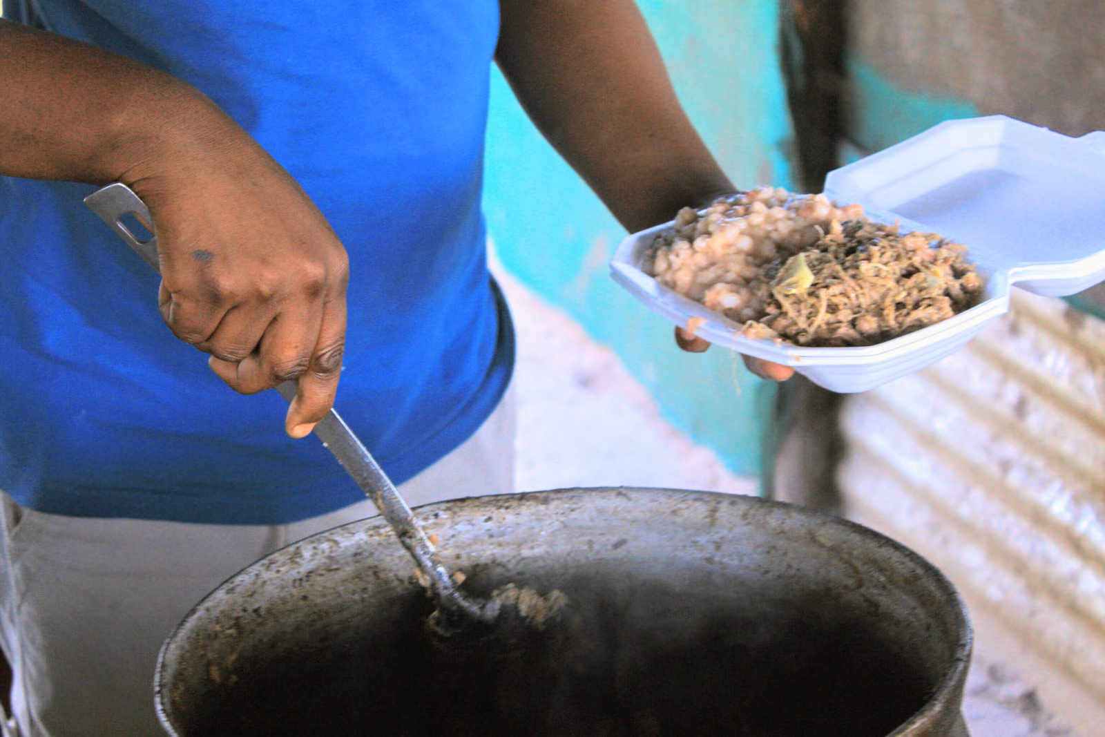 A vendor lifts a spoonful of pale shredded meat from a three-legged cast-iron pot into a waiting container.