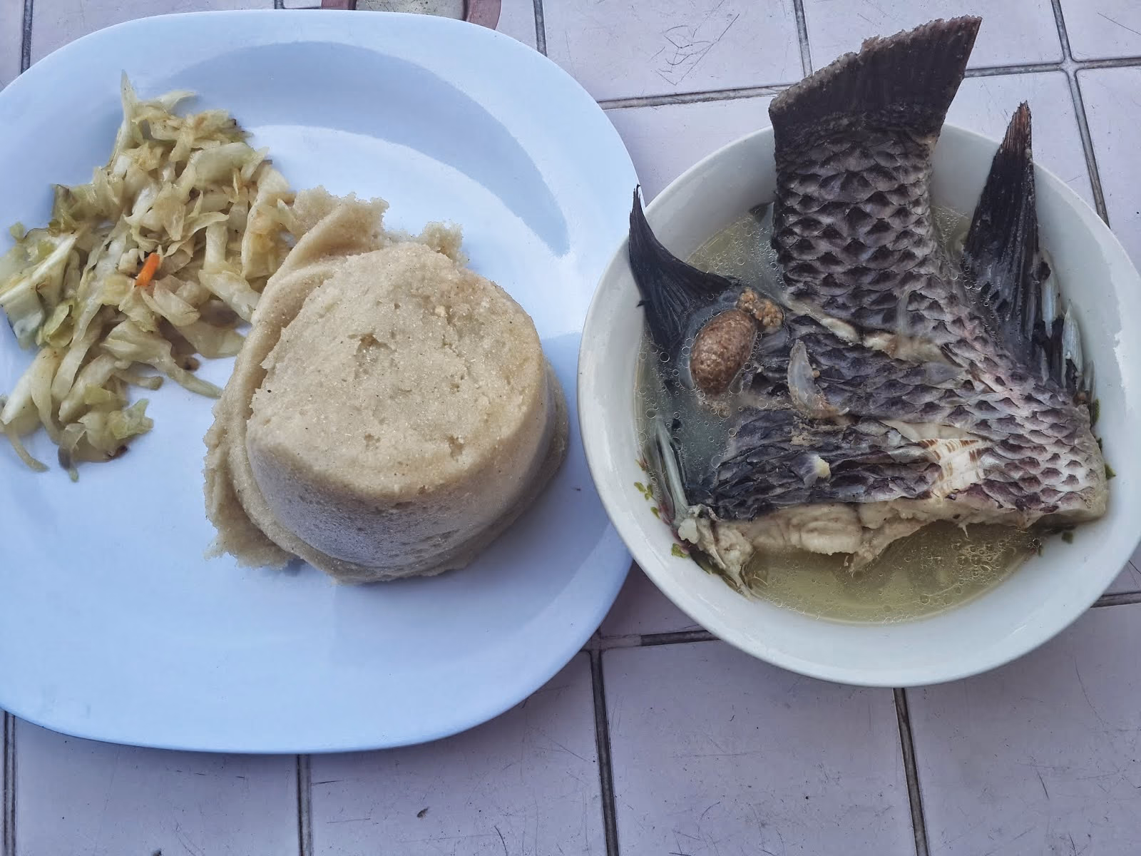 A plate holding a firm white block of ugali beside a whole fried tilapia and a heap of chopped greens.